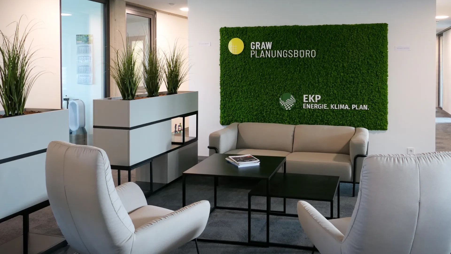 Innenansicht Empfangsbereich Planungsbüro Graw Osnabrück: Lounge mit grünem Moosbild als Verzierung, Logo Graw und EKP, nachhaltige Pflanzen und gemütliche Sitzmöbel – modernes, ökologisches Büro für TGA-Planung und Gebäudetechnik